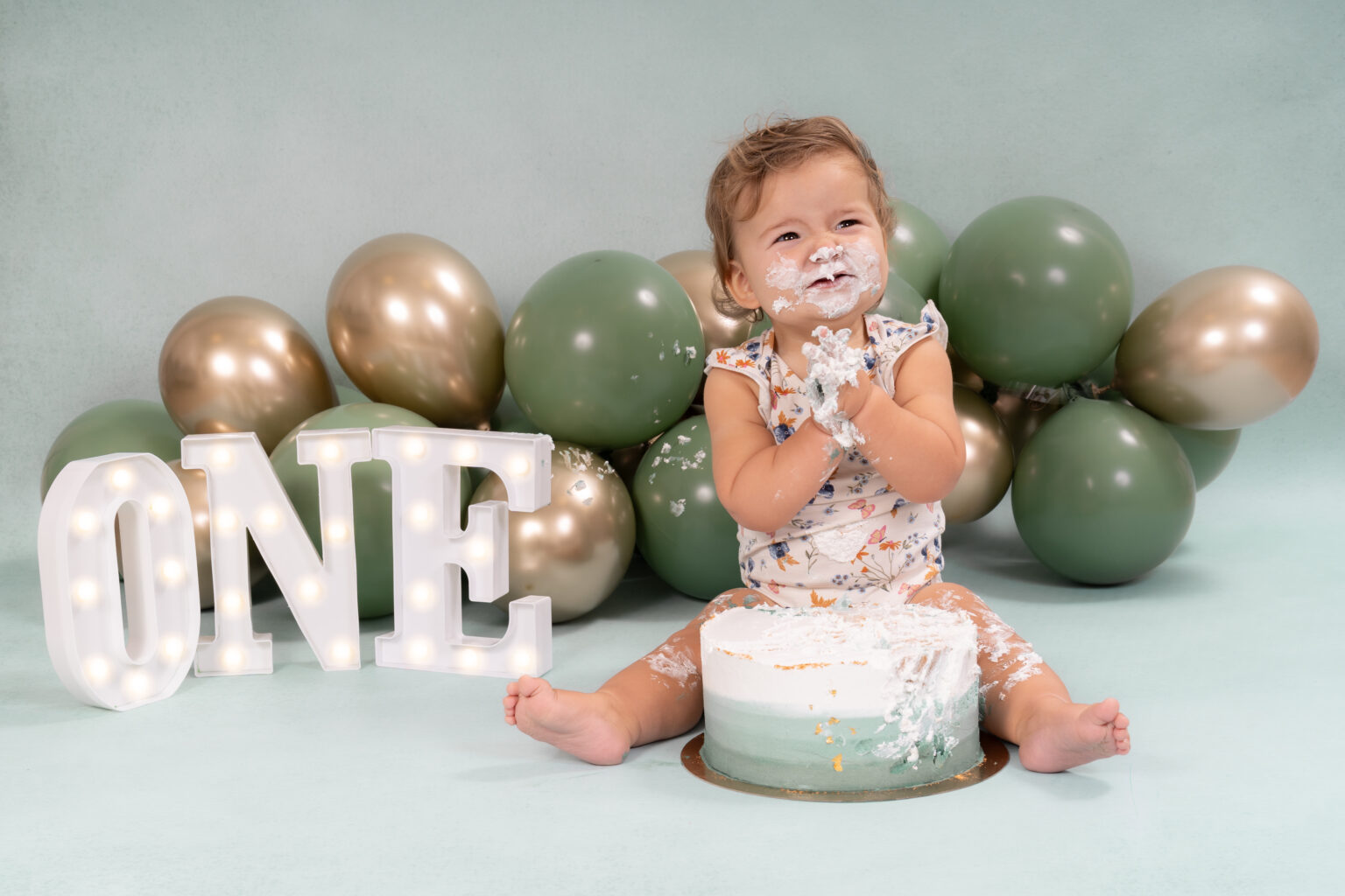Meisje in bloemetjesjurk klapt in haar handen bij een cakesmash met groen en goud