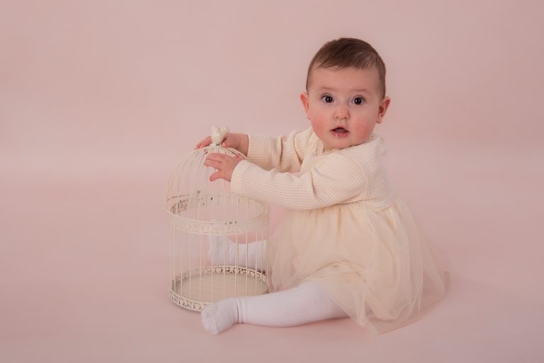 Baby van ongeveer zes maanden zittend bij een vogelkooi tijdens een sittershoot, roze achtergrond