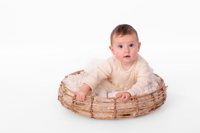 Baby van ongeveer zes maanden zittend bij een rieten mand tijdens een sittershoot, witte achtergrond
