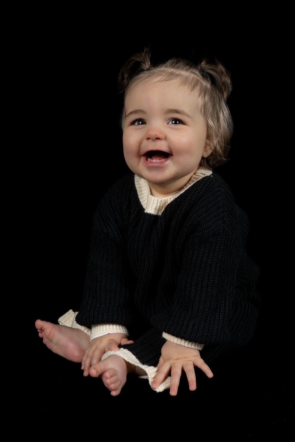 Lachende baby zit zelfstandig in een donkere trui tijdens een sittershoot in de studio